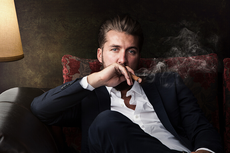 A young man sits in a chair while enjoying a fine cigar.
