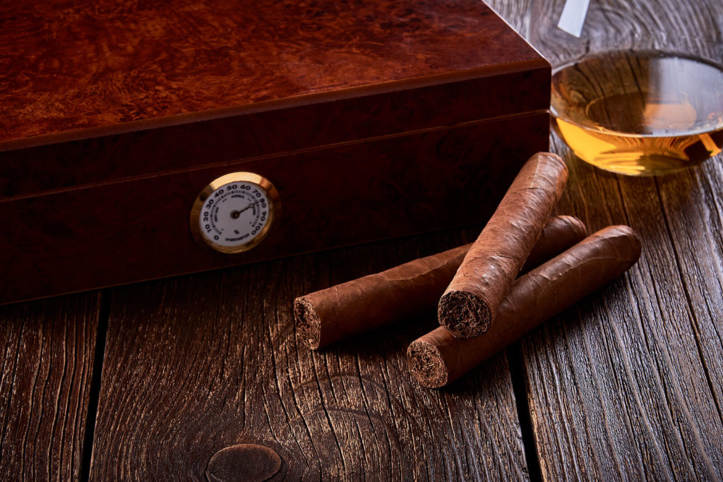 Two cigars sit outside a stylish humidor with a glass of whiskey.